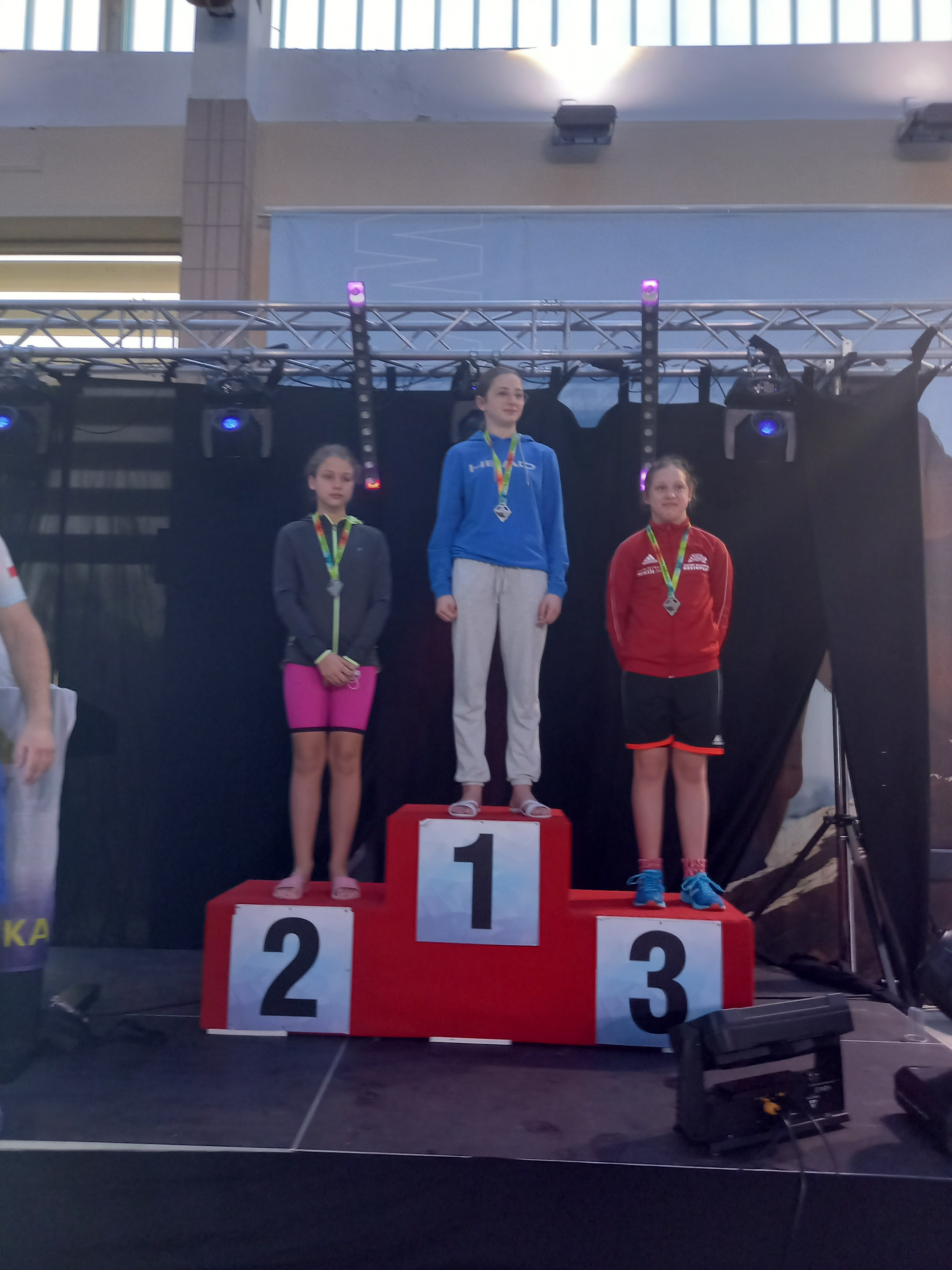 Podium, na którym stoi trzech medalistów. Na miejscu nr 2 (pierwsza z lewej) Karolina Klajmon.