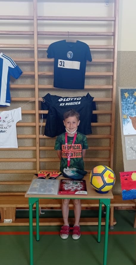 Uczeń stojący przed swoim stoiskiem. Na stoliku znajdują się m.in: piłka nożna, rękawice i buty piłkarskie. Prezentuje swoje hobby związane z piłką nożną.