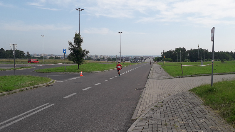Przedstawia Piotra Talika w biegu. Na pierwszym planie, ubranego w czerwoną koszulkę i czarne krótkie spodenki. W tle pozostali biegacze.