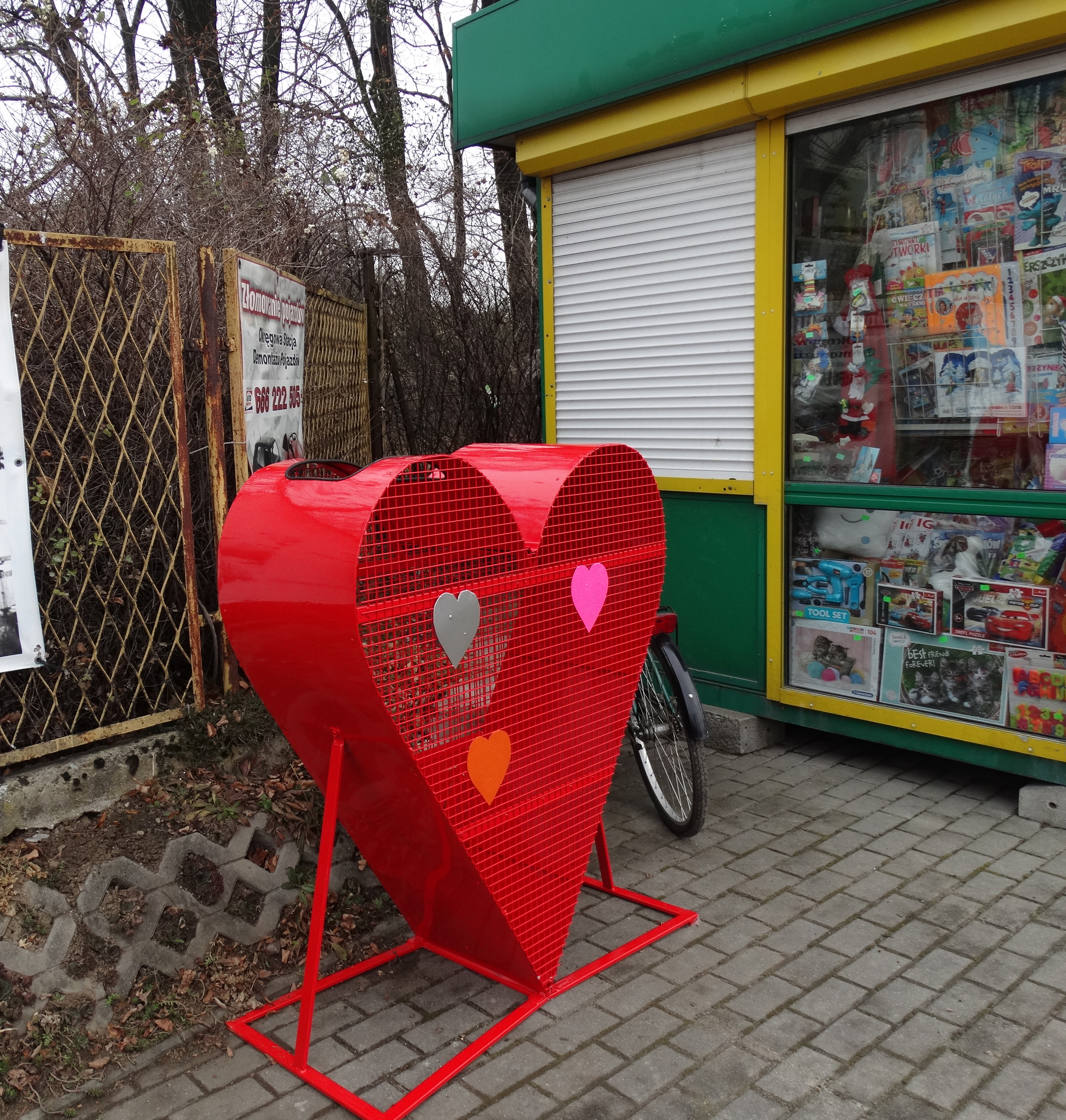Na zdjęciu metalowy kosz na plastikowe nakrętki w kolorze czerwonym, stojący obok kiosku w Rudzicy.
